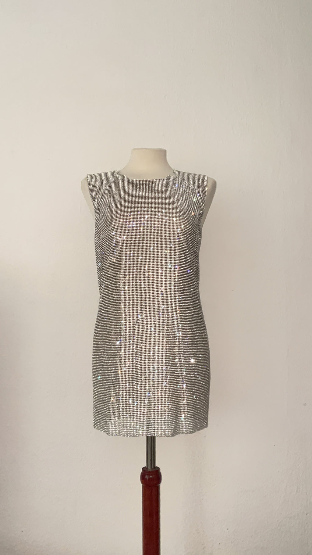 A sleeveless, silver chainmail sequin mini dress. The dress is covered in small, reflective crystal rhinestone, creating a shimmering effect. The dress has a simple, straight cut and appears to be made of a lightweight material. The dress is in the style of Kate Moss’ Versace Dress and Dua Lipa’s bachelorette dress