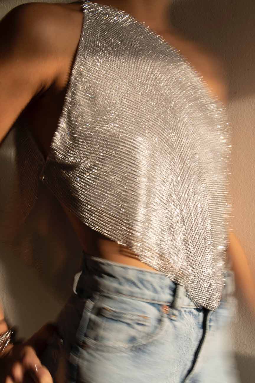 Model wearing a chainmail crystal mesh top in color Silver. One shoulder with silver chains on the side. Embellished rhinestone sequin crop top.
